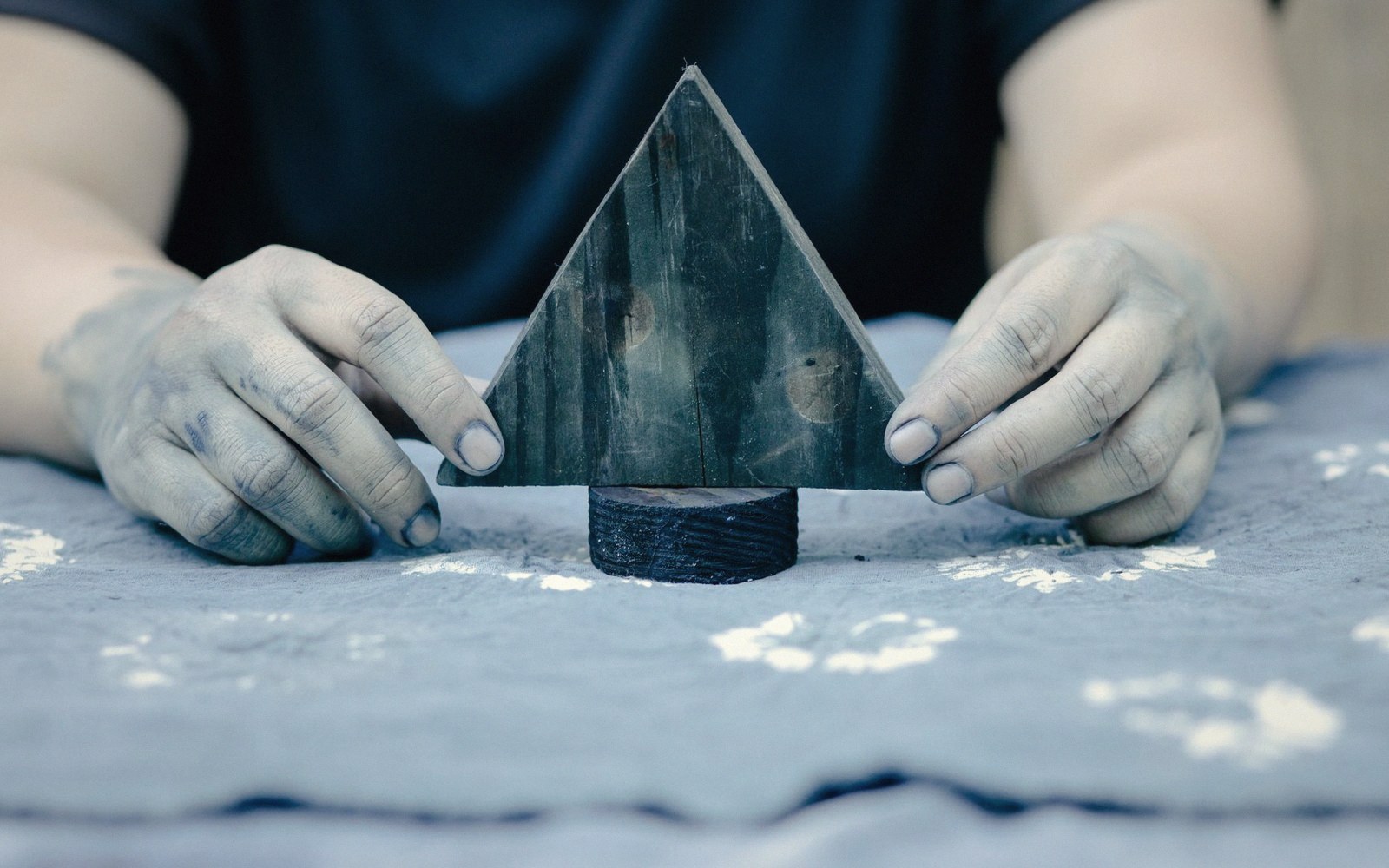Dye-stained hands hold a triangular wooden block stamp upright on cloth